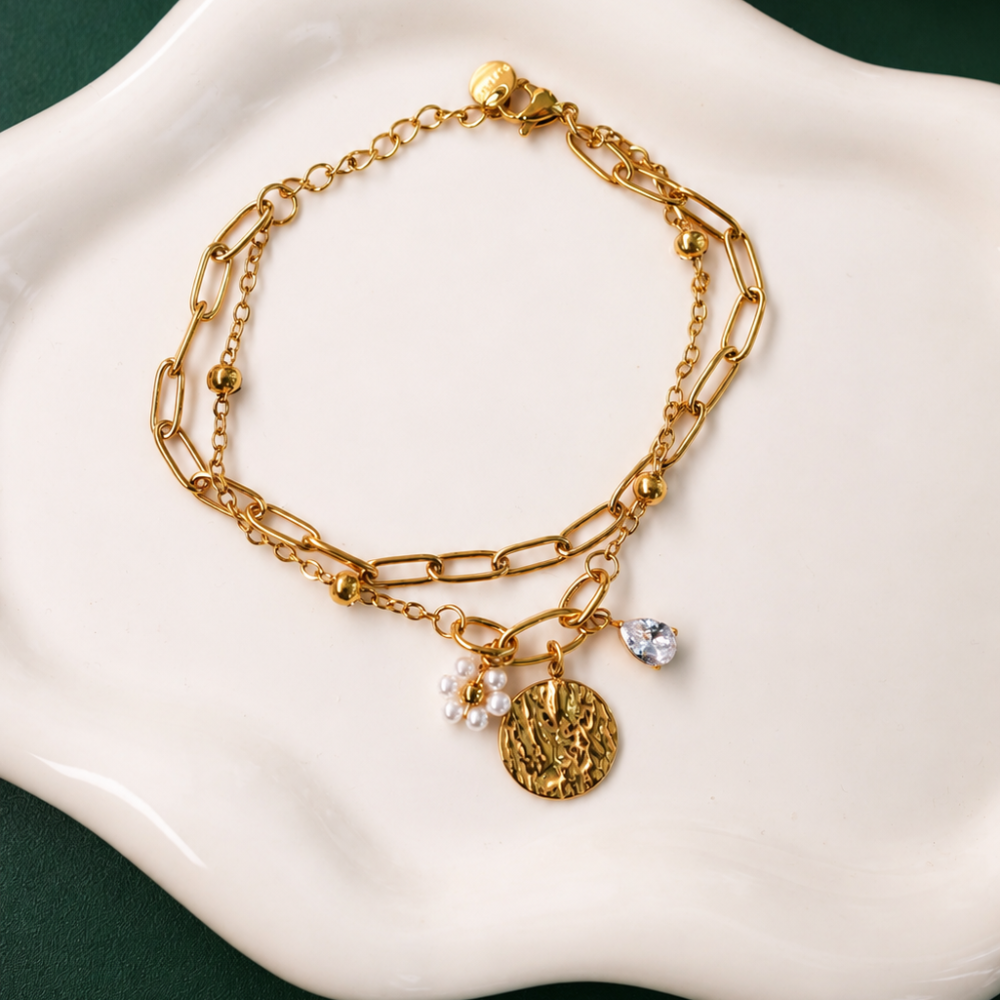 A gold layered bracelet with pearl-style details, a clear stone charm, and a textured pendant. A feminine piece from the Neréa Anthi Curated Collection.
