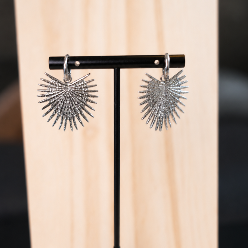 Silver stainless-steel hoop earrings with sculptural sunburst heart pendants. A bold statement pair from the Neréa Anthi Curated Collection.